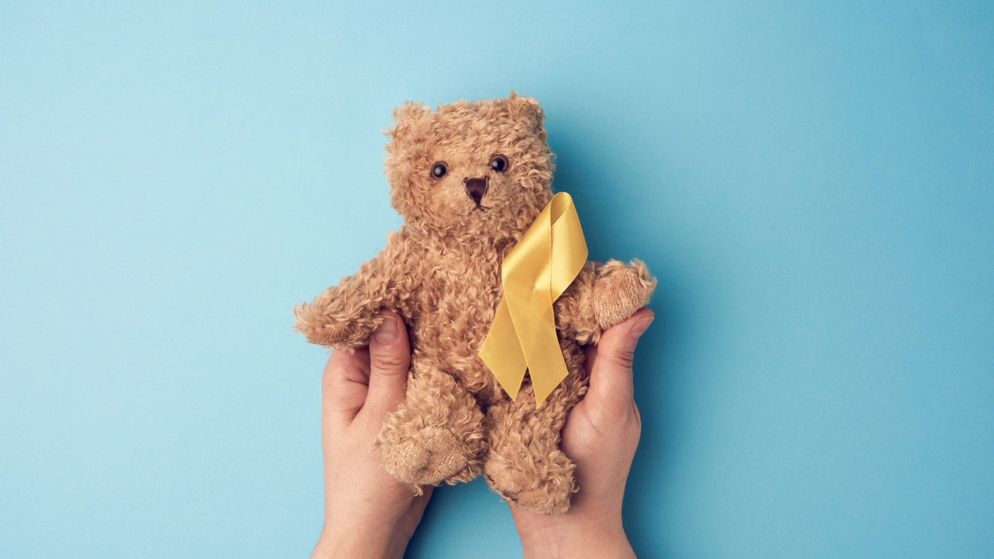 female hands hold a small teddy bear with a yellow ribbon folded in a loop on a blue background. concept of the fight against childhood cancer. problem of suicides