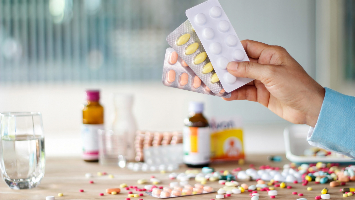 Hand holding medicines pill pack with colorful drugs spread on wooden table in the room background