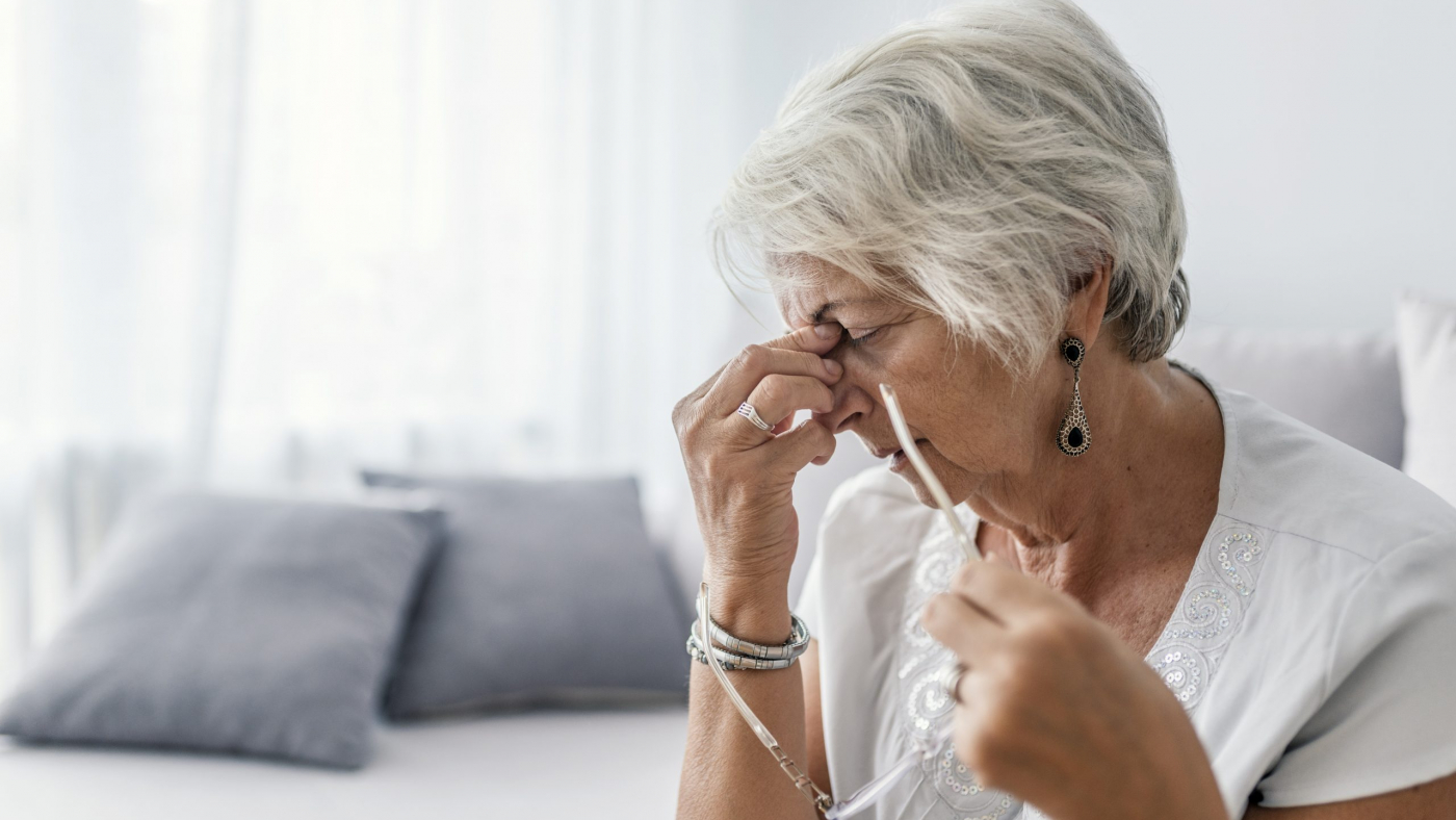 Mature woman sitting on a white sofa in a home touching her head with her hands while having a headache pain and feeling unwell. Senior woman with headache