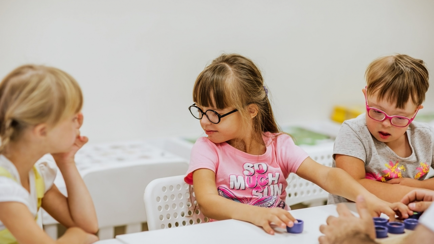 Young kids with down syndrome studying and playing in the room with white desks. Focus is at the girl in the middle.