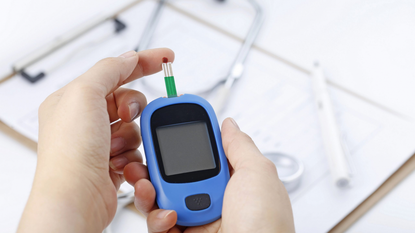 Hand holding a blood glucose meter measuring blood sugar, the background is a stethoscope and chart file