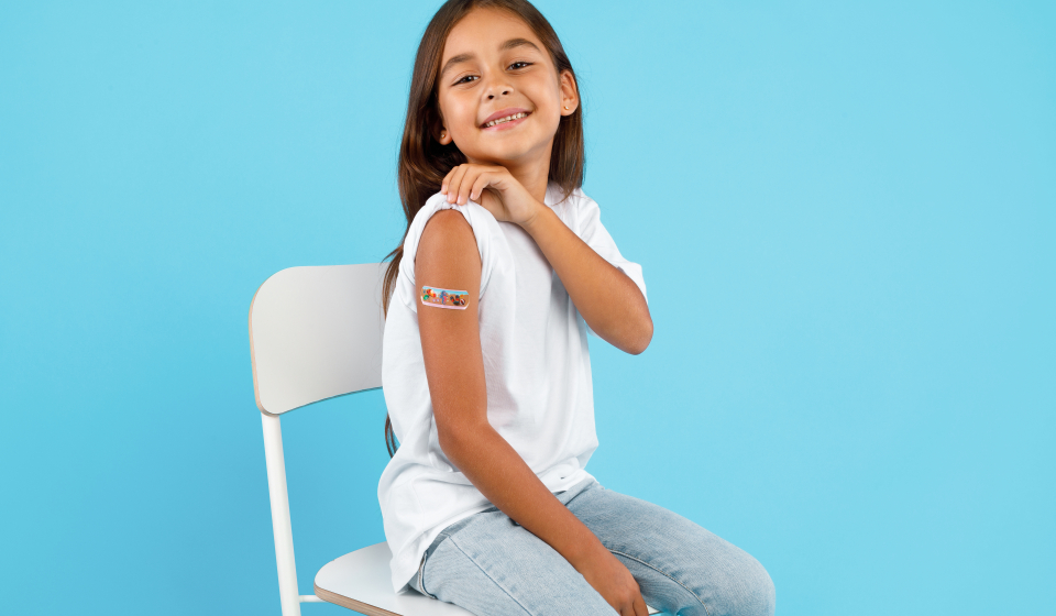 La vacunación de los niños. Happy Vaccinated Kid Girl Showing Arm With Adhesive Bandage After Vaccine Injection Sitting On Blue Studio Background. Niños Y Covid-19 Prevención, Inmunización Antiviral